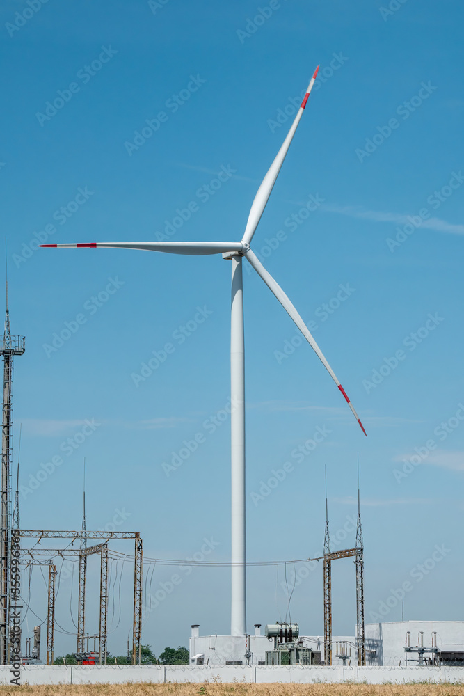 Windmill produces clean energy. Wind turbine generates ecofriendly and renewable energy near electricity distribution substation against blue sky