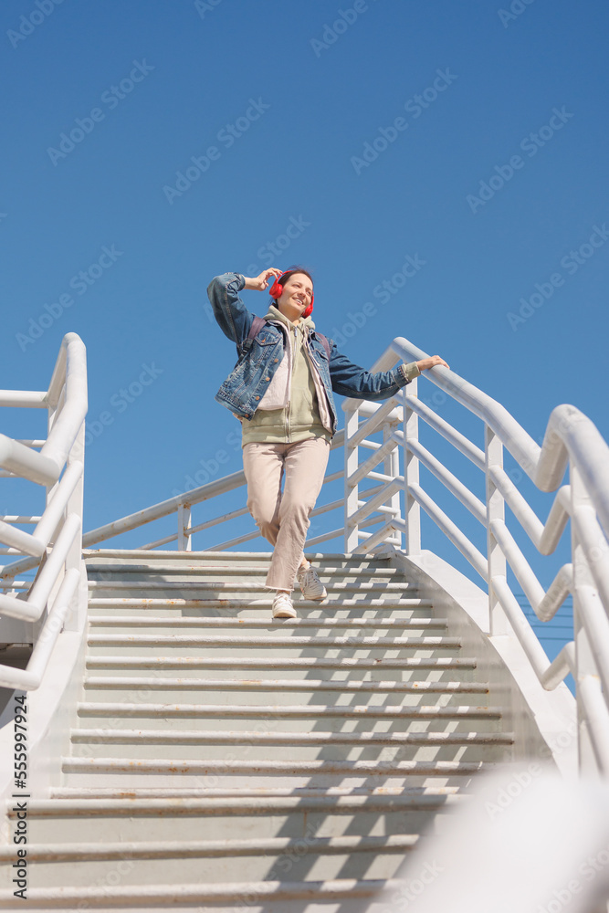 woman dances and sings on stairs, listens to music or podcast with red headphones, full-length. happy lady smiles and spends time alone with herself. take walk through urban places of city.