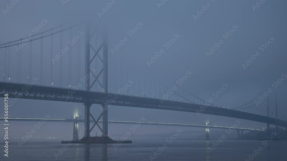 A large suspension bridge over the sea bay covered with fog or haar. The lights of cars moving across the bridge. Video with the panning camera motion. Forth Road Bridge, Scotland, United Kingdom