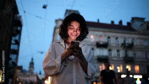 Smiling woman using smartphone on street with night city lights on background