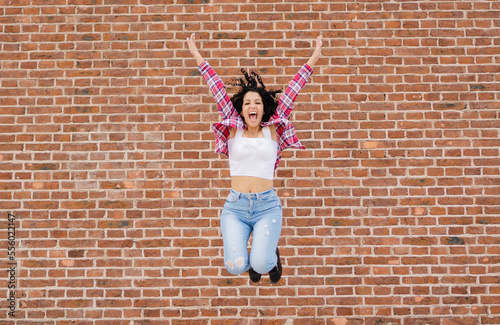 Young latina, hispanic and caucasian woman with curly hair happy jumping with arms up. Brick background. Copy space. © juanpablo