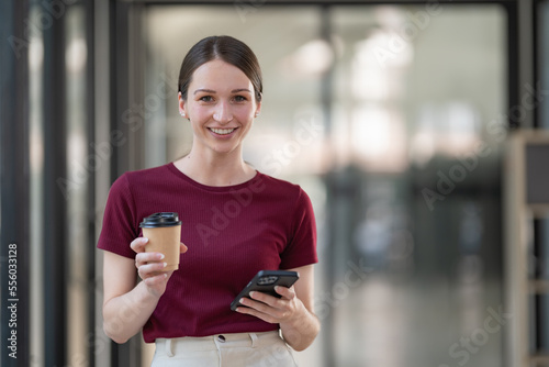 Smiling young businesswoman holding smartphone and coffee cup.
