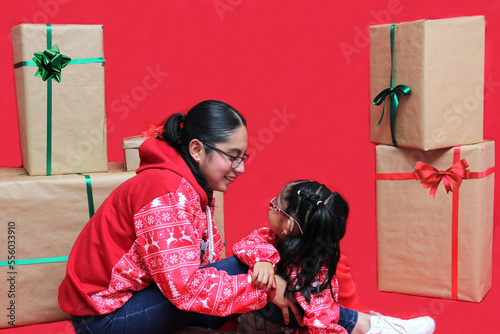 Latina mom and daughter with glasses wear ugly Christmas sweaters and show their love to each other on a red background between large gift boxes with bow
 photo