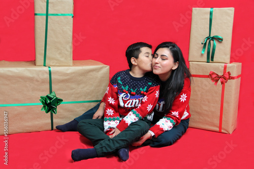 Latina mom and son wear ugly Christmas sweaters and show their love to each other on a red background between large gift boxes with bow photo