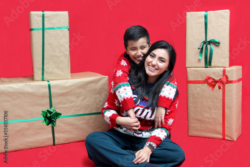 Latina mom and son wear ugly Christmas sweaters and show their love to each other on a red background between large gift boxes with bow photo