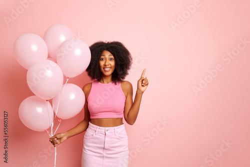 Horizontal shot of curly haired young woman, shows index finger aside on a blank space, poses with pink balloons in a hand, dressed in pink shirt and top, isolated over pink background