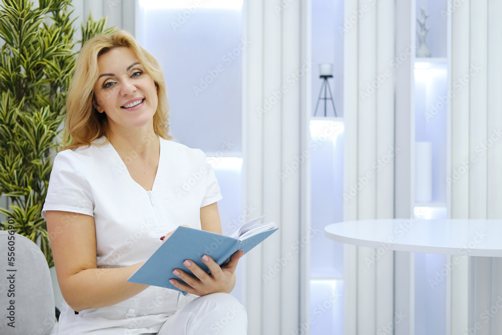 Beautiful mature female cosmetologist in a medical suit sits in a chair, makes notes in her notebook, smiles and looks at the camera Beautiful mature female cosmetologist in a medical suit sits in a chair, makes notes in her notebook, smiles and looks at the camera