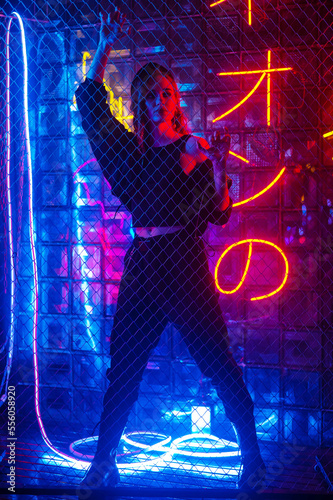 Caucasian woman in neon studio behind chain-link mesh. 