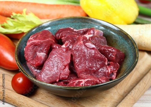 Raw deer meat for venison ragout or goulash. Bowl with pieces of deer meat on cutting board, fresh vegetable for cooking around.