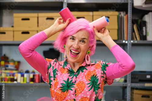 Dressmaker with pink hair and colorfull clothes making funny faces with sewing thread reels in a sewing workshop.