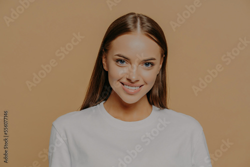 Portrait of beautiful woman with long dark straight hair smiles gently wears casual white jumper has minimal makeup poses against brown background. Women beauty and positive emotions concept