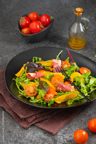 Fresh salad with arugula, cherry tomatoes, orange slices and smoked duck slices on a black plate on a dark concrete background. Served with olive oil dressing. Salad recipes.