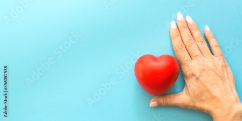 Fototapeta Naklejka Na Ścianę i Meble -  a female hand protects a red toy heart on a blue background, a banner on the left copyspace, children's health insurance, a woman's love, save a life, take care of a woman,