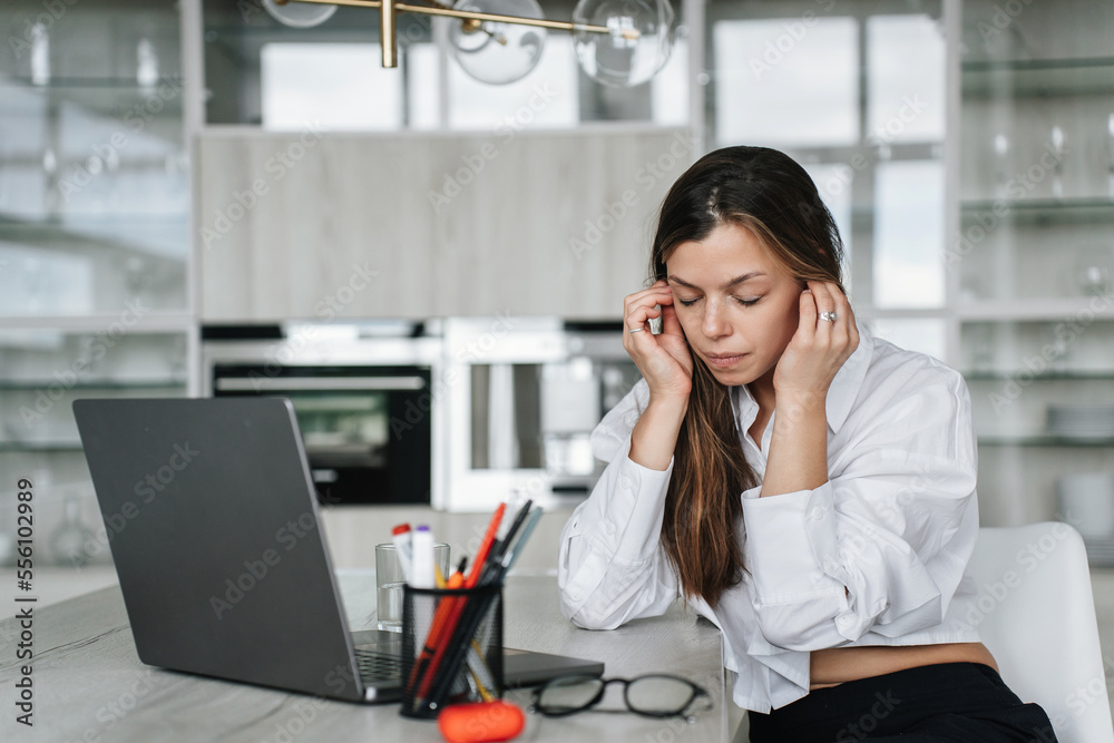Exhausted young woman in white shirt and black pants sitting at desk using laptop touches temples feels headache, fatigue tired after hard working at home. Overloaded employee distant working.