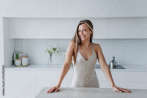 Playful sexy caucasian woman in nightgown standing at kitchen leans on table looks at camera toothy smiles enjoys Sunday morning. Successful model satisfied by new home. Gorgeous lady with long hair.