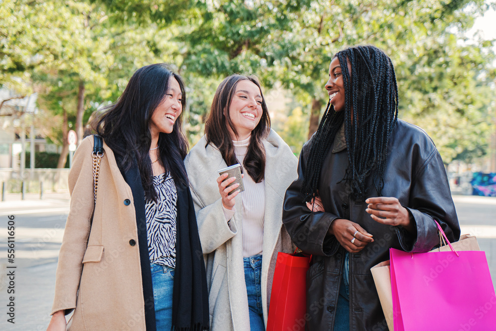 Three multiracial happy young women walking and talking on a shopping day. Group of girls carrying bags smiling on the street on a sale week. Consumism concept. High quality photo Three multiracial happy young women walking and talking on a shopping day. Group of girls carrying bags smiling on the street on a sale week. Consumism concept. High quality photo