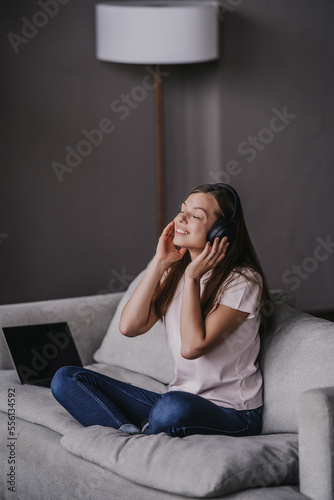 Cheerful hispanic brunette girl in headphones eyes closed sits on sofa listening music at home. Young italian pretty woman in white t-shirt relaxing after hard studying. Youth and entertainment.