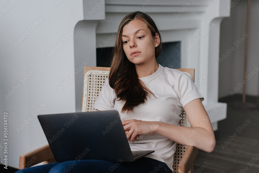 Fototapeta premium Focused Hispanic young woman in casual clothes sitting on chair with laptop at home against fireplace at home. Caucasian attractive female entrepreneur remote working, using computer. Purposeful girl.