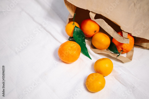 Beautiful ripe tangerines with leaves fall out of a brown paper bag on a white crumpled blanket. Minimalism, health, aesthetics.