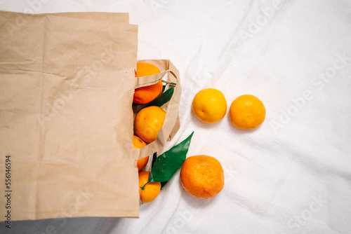 Beautiful ripe tangerines with leaves fall out of a brown paper bag on a white crumpled blanket. Minimalism, health, aesthetics.