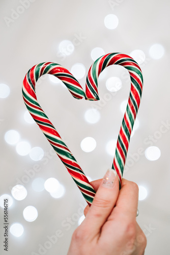 A woman's hand holds two Christmas candy lollipops creating a heart shape. There is a blurred garland on the background, lights