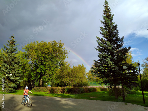 Kazan, Tatarstan. August 23, 2020. Raduai after the rain in the forest nature. Tree crown and rainbow. A boy in a mask rides a bicycle in the park.