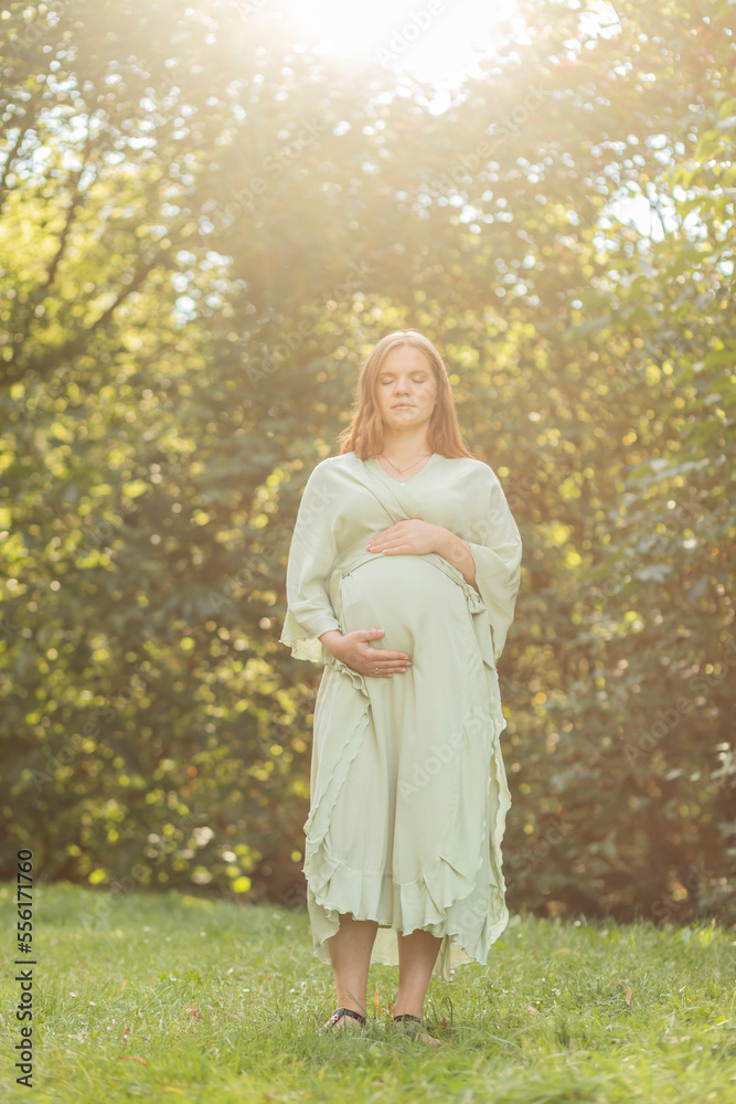 Young pregnant woman puts hands on big belly, green trees,sky on background. Brown haired female wears dress. Magic happy pregnancy. Child delivery preparation,emotional connection with baby. Vertical Young pregnant woman puts hands on big belly, green trees,sky on background. Brown haired female wears dress. Magic happy pregnancy. Child delivery preparation,emotional connection with baby. Vertical