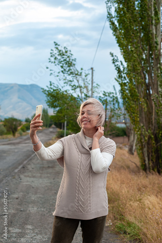 a young adult woman takes a selfie photo of herself with a mobile phone stretched out and held in her hand against the backdrop of a beautiful scenic nature in a mountainous area at sunset