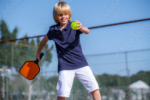 Happy blonde boy playing pickleball game, hitting pickleball yellow ball with paddle, outdoor sport leisure kids activity. photo