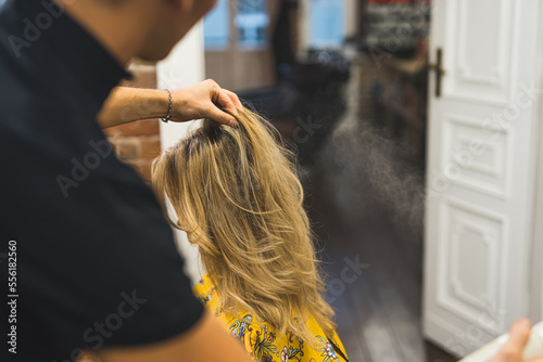 The process of styling with hairspray blond female client's hair by unrecognizable blurred hairdresser. Medium close-up shot. High quality photo