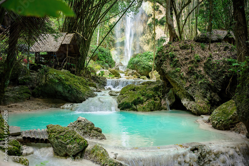 A clearing in the tropical jungle with a turquoise pond in the foreground and many moss-covered rocks. In the background is a large waterfall and many trees