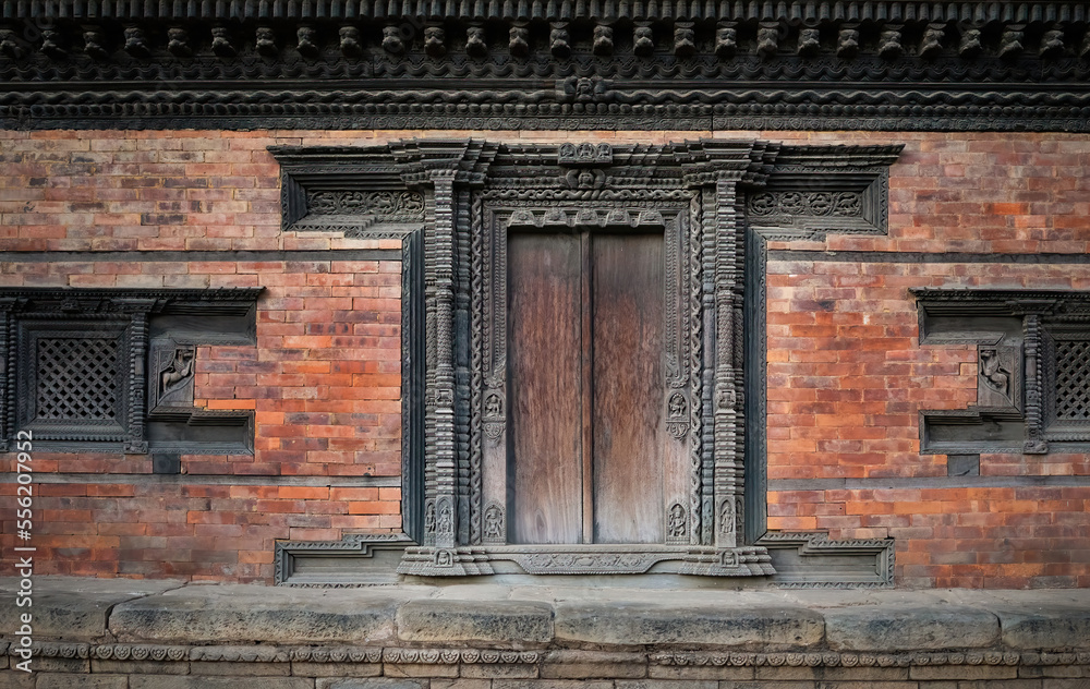 Frontal view of a traditional Nepalese temple facade with red bricks and ornamental carvings. A closed wooden door made of dark wood and two closed windows