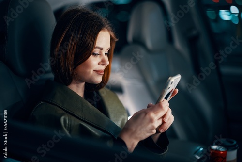 a close horizontal portrait of a stylish, luxurious woman in a leather coat sitting in a black car at night in the passenger seat, looking pleasantly into the camera holding a smartphone in her hand © SHOTPRIME STUDIO