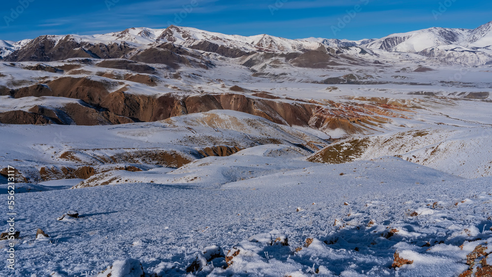 Snow-capped mountains with red-brown slopes against a blue sky. In the foreground is a plateau with dry grass covered with hoarfrost. Altai Mars Kyzyl Chin