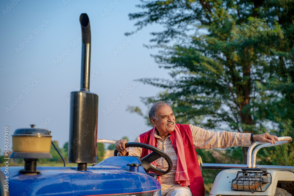 Indian senior farmer sitting on tractor at agriculture field.