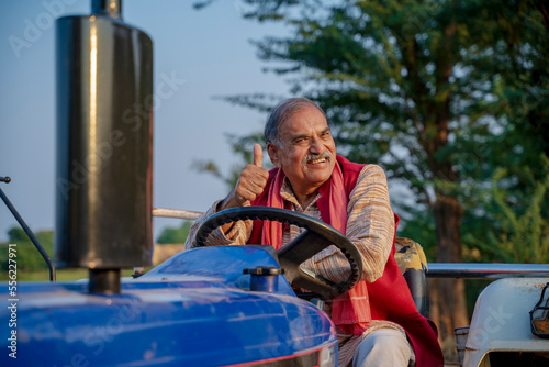 Indian senior farmer sitting on tractor and showing thumps up at agriculture field.