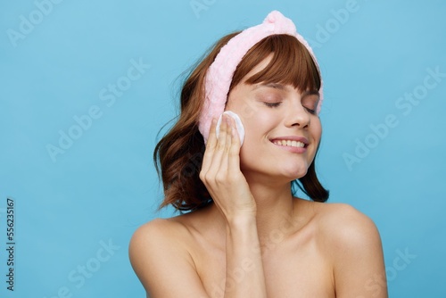 a girl with dark hair stands on a blue background in a white towel with a pink bandage on her head after a bath, removes makeup from her face with a cotton pad. Horizontal studio photography.