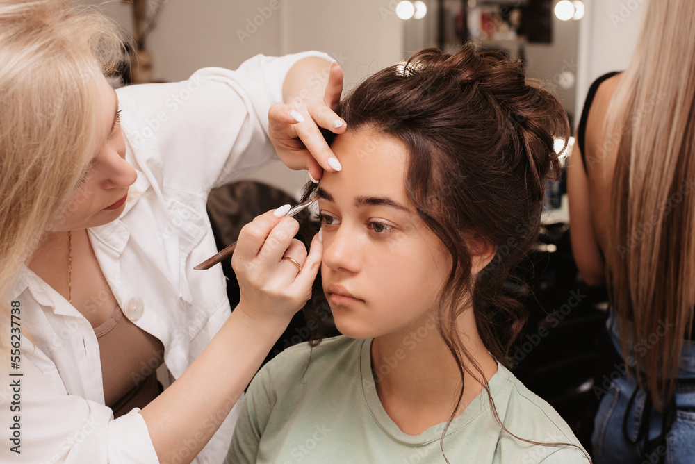 Shooting in a beauty salon. A makeup artist does eyebrow styling for a young dark-haired girl.
