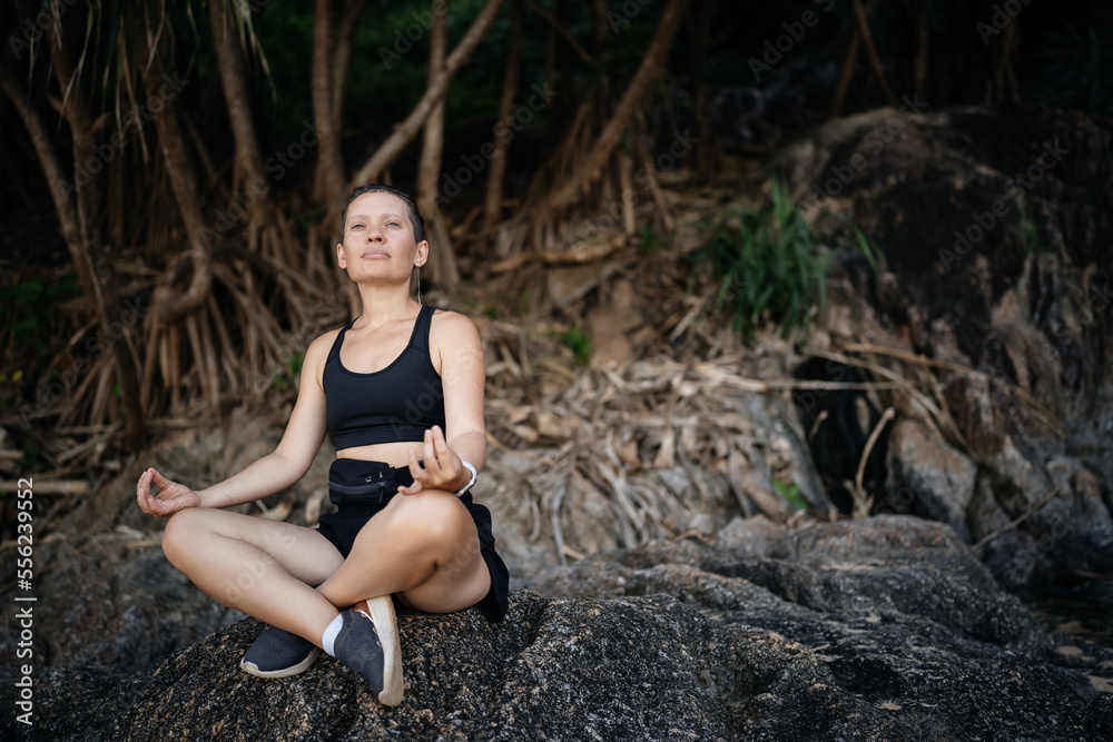 A woman in a black sports top and shorts sitting on the rocks in the tropics is engaged in sports breathing. A woman in a black sports top and shorts sitting on the rocks in the tropics is engaged in sports breathing.