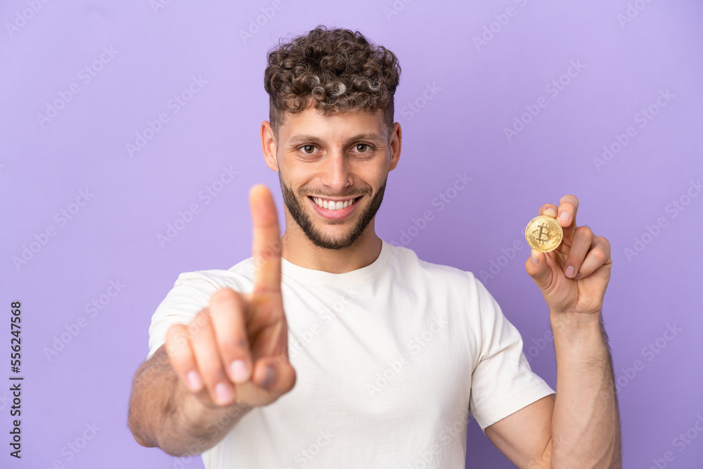 Young caucasian man holding a Bitcoin isolated on purple background showing and lifting a finger Young caucasian man holding a Bitcoin isolated on purple background showing and lifting a finger