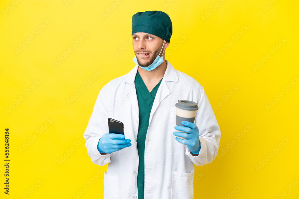Surgeon blonde man in green uniform isolated on yellow background holding coffee to take away and a mobile while thinking something Surgeon blonde man in green uniform isolated on yellow background holding coffee to take away and a mobile while thinking something