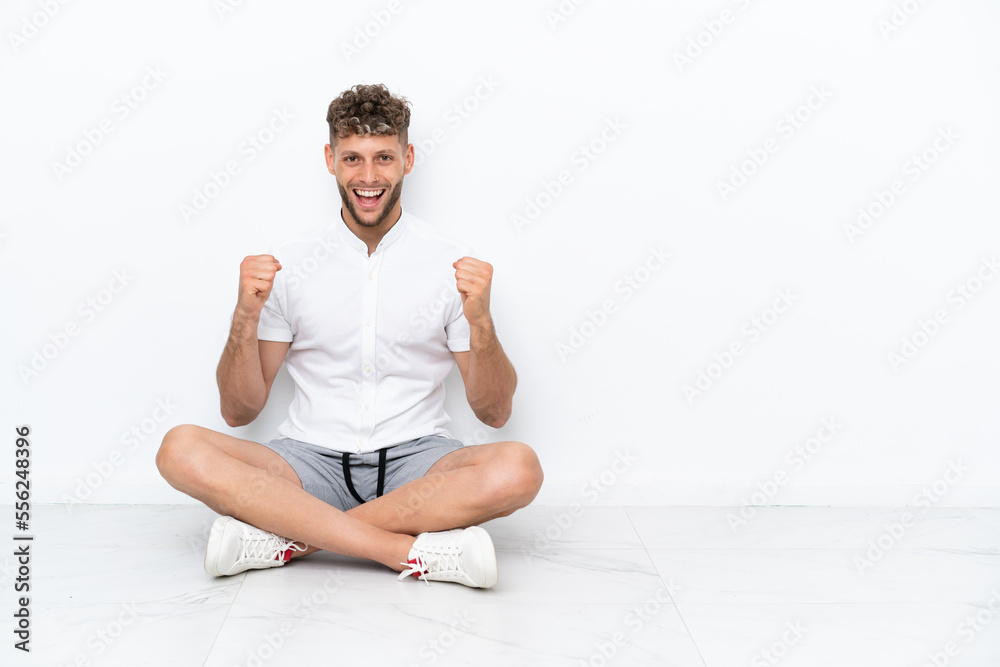 Young blonde man sitting on the floor isolated on white background celebrating a victory in winner position Young blonde man sitting on the floor isolated on white background celebrating a victory in winner position