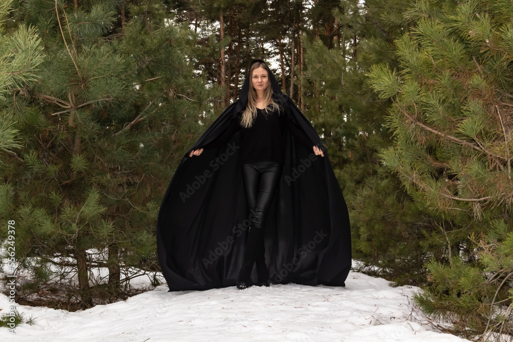 Blonde woman in a black cloak with a hood in winter in a pine forest