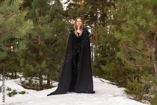 Blonde woman in a black cloak with a hood in winter in a pine forest