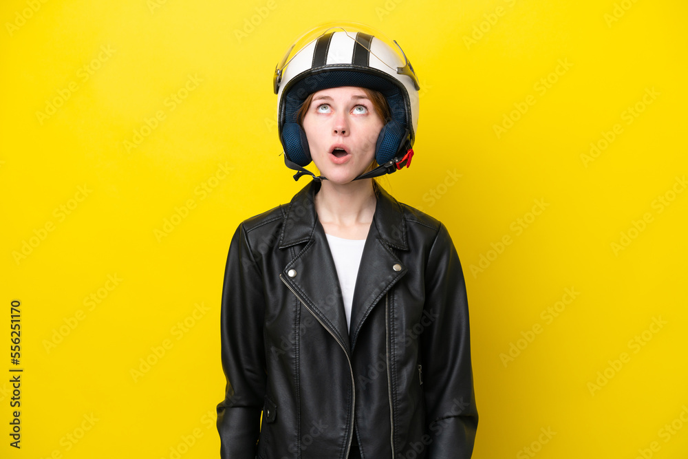 Young English woman with a motorcycle helmet isolated on yellow background looking up and with surprised expression Young English woman with a motorcycle helmet isolated on yellow background looking up and with surprised expression
