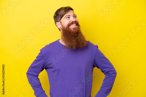 Redhead man with beard isolated on yellow background posing with arms at hip and smiling