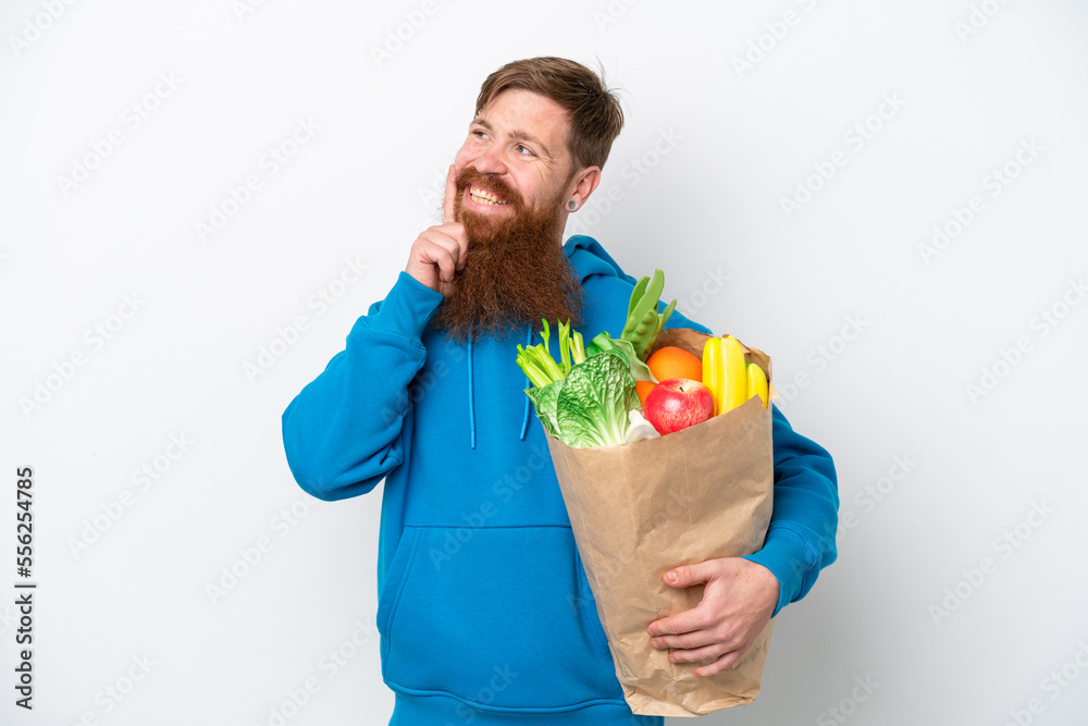 Redhead man with beard holding a grocery shopping bag isolated on white background thinking an idea while looking up Redhead man with beard holding a grocery shopping bag isolated on white background thinking an idea while looking up