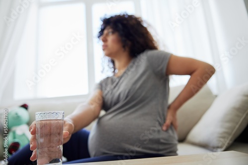 A pregnant woman sits on the couch with lower back pain and headache, strain on the spine during pregnancy drinks water. Lifestyle preparation for childbirth, last month of pregnancy