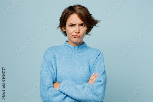 Young sad offended unhappy caucasian woman wearing knitted sweater look camera hold hands crossed folded isolated on plain pastel light blue cyan background studio portrait. People lifestyle concept.