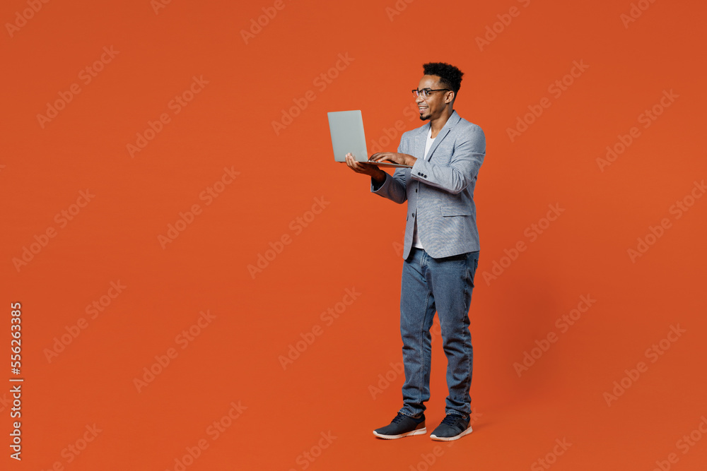 Full body side view fun young employee IT business man corporate lawyer wear formal grey suit shirt glasses work in office hold use laptop pc computer isolated on plain red orange background studio.